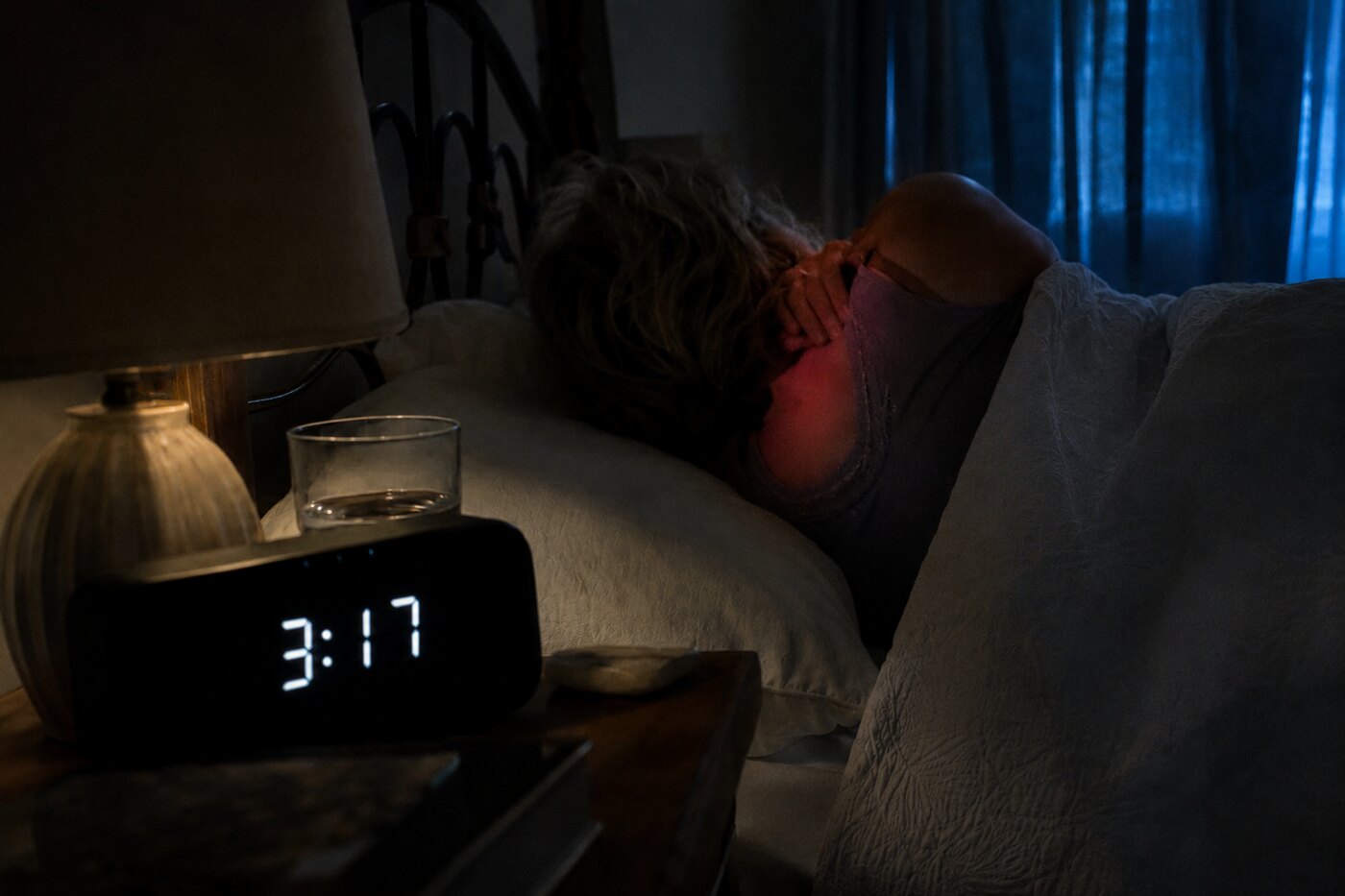 A woman in her 50s awake in bed at 3:17am, a warm glow at her neck, a bedside clock reading 3:17