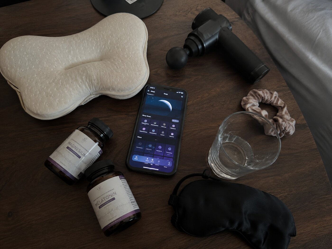 A nightstand crowded with a memory-foam pillow, magnesium, melatonin, a sleep-app phone, a massage gun and an eye mask